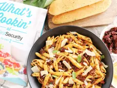 Makaron penne z suszonymi pomidorami, parmezanem i bazylią w czarnej misce, obok pieczywa i książki kucharskiej.