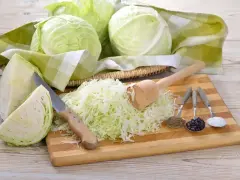 Przygotowanie kiszonej kapusty: ile soli na 10kg? Na desce poszatkowana kapusta, nóż, tłuczek, kminek, pieprz i sól.