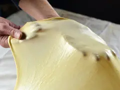 Mani che stendono la pasta matta per una ricetta tradizionale. La pasta è sottile e traslucida, con le dita che creano delle leggere ondulazioni.