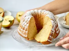 Ein saftiger Kuchen mit Apfelmus, bestäubt mit Puderzucker, daneben Äpfel und ein Glas Apfelmus.