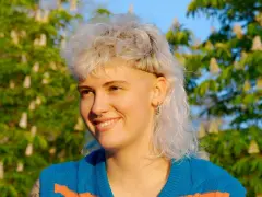 Persona sonriente con un moderno mullet rubio, aretes y un suéter azul con rayas naranjas, frente a un árbol en flor.