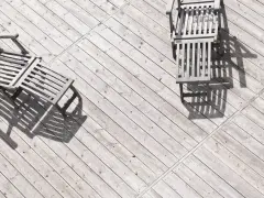 Tre solstolar i trä står på en sliten altan. Solen kastar långa skuggor över siox trall.