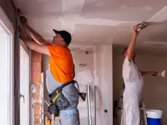 Deux ouvriers rénovent un bâtiment, l'un ajuste des câbles près d'une fenêtre, l'autre enduit le plafond. Ces travaux sont essentiels pour les aides aux collectivités locales.