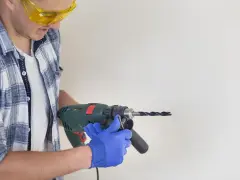 Man in safety glasses and blue gloves holding a drill to a white surface.