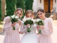 Trzy uśmiechnięte kobiety z bukietami kwiatów. Panna młoda w białej sukni i dwie druchny w różowych sukienkach.