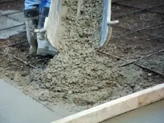 Wylewanie świeżego betonu na zbrojenie. Trwa naprawa betonu, widać buty pracownika i metalowe pręty.