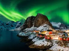 Grön norrsken dansar över snöklädda berg och röda fiskestugor vid en vik i Norge.