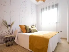 Habitación con papel tapiz de ramas y gotele moderno. Cama con cojines verdes y naranjas, y manta mostaza.