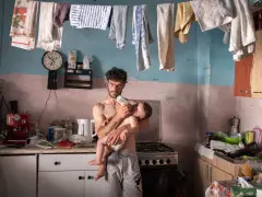 Emiliano Dante, padre single, nutre il suo bambino in una cucina disordinata, con panni stesi ad asciugare.