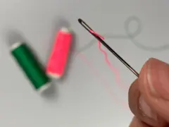 Dedos sujetan una aguja para meter hilos, con carretes de hilo verde y rosa desenfocados al fondo.