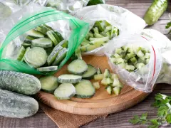 Zamrożone ogórki w plastrach i kostkach w woreczkach, obok świeżych ogórków i natki pietruszki.