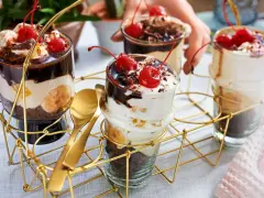 Schokoladige Dessert-Rezepte in Gläsern mit Bananen, Sahne und Kirschen. Goldene Löffel liegen bereit.