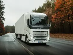 Biała ciężarówka Volvo FH12 z naczepą jedzie po mokrej drodze wśród jesiennego lasu.