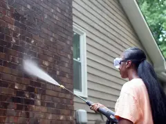 Woman pressure washing a dark acid wash brick wall, restoring its natural beauty.