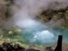 Gorący, bulgoczący onsen otoczony skalistym, parującym krajobrazem. Woda ma turkusowy odcień, a brzegi pokryte są żółtymi osadami.