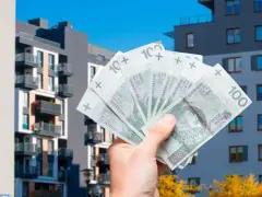 Dłoń trzyma banknoty 100 zł na tle nowoczesnych budynków mieszkalnych. Symbolizuje to podatek od sprzedaży nieruchomości.