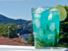 Orzeźwiający drink z blue curacao i sprite, z miętą i lodem, ozdobiony plasterkiem limonki, na tle zielonych wzgórz.