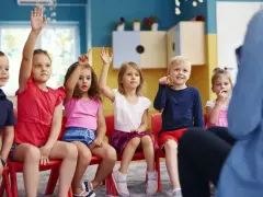 Dzieci na czerwonych krzesłach podnoszą ręce, jakby odpowiadały na pytanie, czy zerówka jest obowiązkowa.