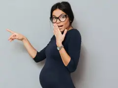Donna in gravidanza con occhiali indica sorpresa, coprendosi la bocca con una mano.