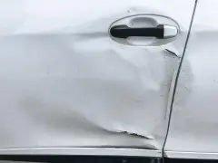Widoczne wgniecenie i zarysowania na srebrnych drzwiach samochodu, świadczące o uderzeniu drzwiami na parkingu.