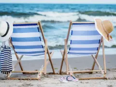 Dwa leżaki na plaży, gotowe na idealny weekend nad morzem. W tle fale, a obok kapelusze i torba plażowa.