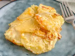 Ein Stück Kartoffelgratin einfach mit Sahne, goldbraun gebacken, auf einem blauen Teller. Daneben eine Gabel.