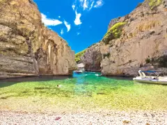 Spiaggia di ciottoli e acque cristalline tra le rocce dell'isola di Lissa, con persone che nuotano e barche.