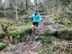 Kvinna springer i skogen, fokuserad på att springa marathon. Mossbeklädda stenar och träd skapar en utmanande terräng.