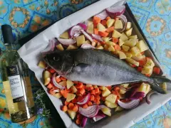 Ricciola al forno pronta per essere infornata con patate, carote e cipolle rosse. Accanto, una bottiglia di vino bianco.