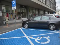 Niebieskie miejsce parkingowe dla niepełnosprawnych z białym symbolem wózka inwalidzkiego. Obok stoi ciemny samochód.