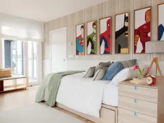Habitación adolescente chico con cuadros de superhéroes, cama blanca con mantas verdes y cojines, mesita de noche de madera y ventana grande.