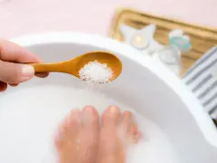 A hand adds epsom salt and baking soda to a foot soak, with feet submerged in the milky water.