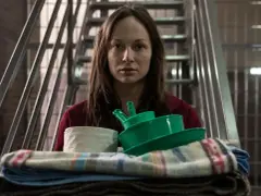 Pati 2, zamyślona, niesie stos rzeczy: zielone miski, papier toaletowy i koce. Za nią metalowe schody i kraty.