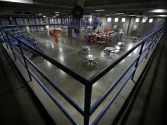 Wnętrze więzienia w USA. Więźniowie w pomarańczowych strojach poruszają się po betonowym dziedzińcu, gdzie znajdują się stoły i łóżka.