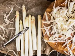 Frischer weißer Spargel wird geschält, die Spargelschalen liegen daneben. Ein Sparschäler liegt auf den Spargelstangen.