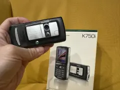 Dłoń trzyma czarny telefon Sony Ericsson K750i, obok stoi pudełko z tym modelem.
