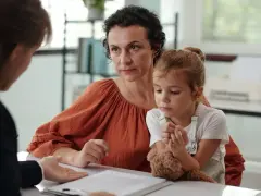 Matka z córką rozmawiają z urzędniczką o tym, ile wynosi renta na autyzm. Dziecko trzyma misia.