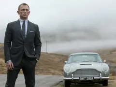Uomo con impeccabile 007 outfit, completo gessato e cravatta, posa accanto a una Aston Martin d'epoca in un paesaggio nebbioso.