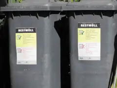Dwa szare kosze na śmieci z etykietami "REST MÜLL". Etykiety zawierają informacje o tym, co można, a czego nie można wyrzucać. Wymiary kosza na śmieci są standardowe.