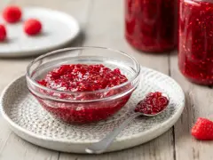 Una ciotola di vetro piena di marmellata di lamponi fatta in casa, con un cucchiaino e lamponi freschi sullo sfondo.