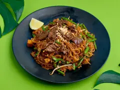 Pyszne pad thai z wołowiną, fasolką szparagową i orzeszkami na czarnym talerzu, ozdobione ćwiartką cytryny i liśćmi monstery.