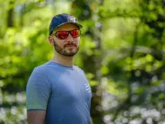 Mężczyzna w czapce i sportowych okularach przeciwsłonecznych, z odbiciem lasu w soczewkach.