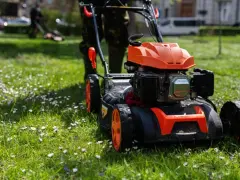Pomarańczowa kosiarka spalinowa pchana z czarnym silnikiem na zielonej trawie z kwiatkami.