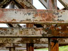 Metalowe belki konstrukcji z widoczną łuszczącą się farbą i rdzą. Postępująca korozja niszczy stal, tworząc rdzawy nalot i ubytki.
