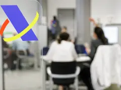 Logo de Hacienda en primer plano. Al fondo, personas atendiendo trámites, quizás esperando cuando devuelve Hacienda.