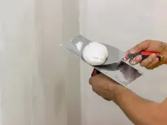 Mani che usano spatole per applicare stucco su un muro. Lo stucco serve a livellare e riparare le imperfezioni.