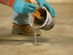 Man con guantes azules vierte hormigón líquido de un cubo para reparar el suelo. Aprende como hacer hormigón para suelo.