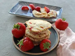 Ein Stapel Erdbeer-Pancakes mit frischen Erdbeeren auf einem Teller. Ein weiterer Teller im Hintergrund zeigt geschnittene Erdbeeren und Pancakes mit Erdnussbutter.