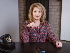 Kobieta w kolorowej bluzce siedzi przy biurku z telefonem i okularami.