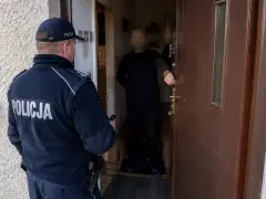 Policjant w mundurze z napisem POLICJA stoi przed drzwiami, gdzie inny mężczyzna otwiera wejście. Sytuacja może być związana z art. 221 kk.